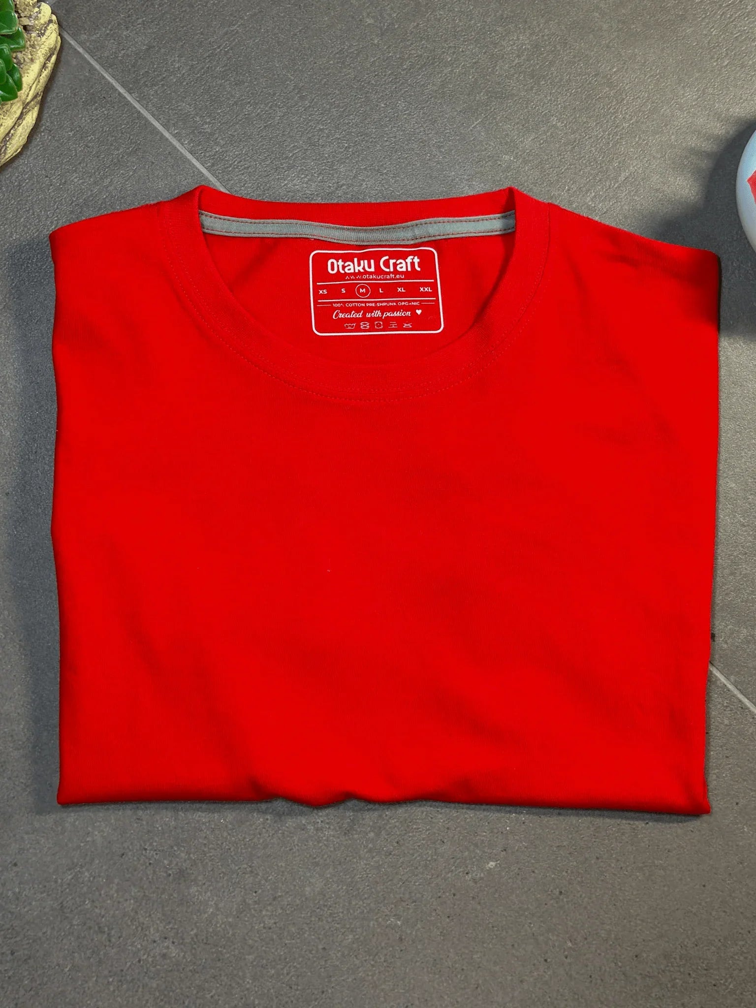 Maglietta rossa unisex in cotone piegata su sfondo grigio, etichetta visibile al collo.