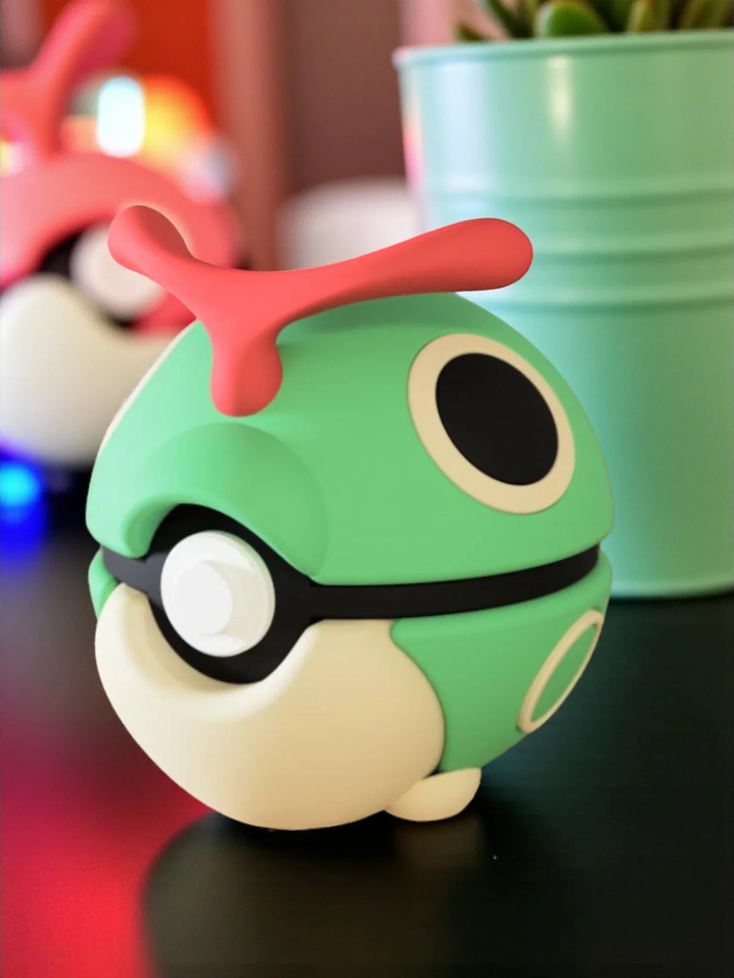 Pokeball decorativa ispirata a Caterpie, verde con dettagli rossi e base magnetica, su tavolo nero