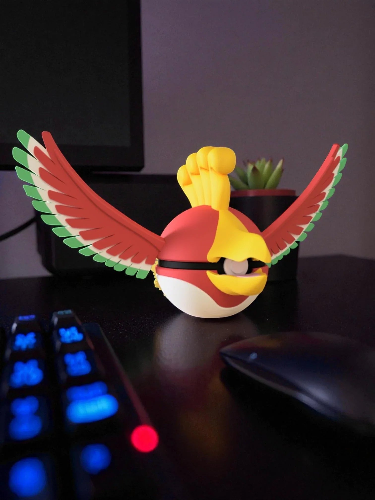 Pokeball ispirata a Ho-Oh colorata, con ali aperte, su scrivania nera vicino mouse e tastiera.