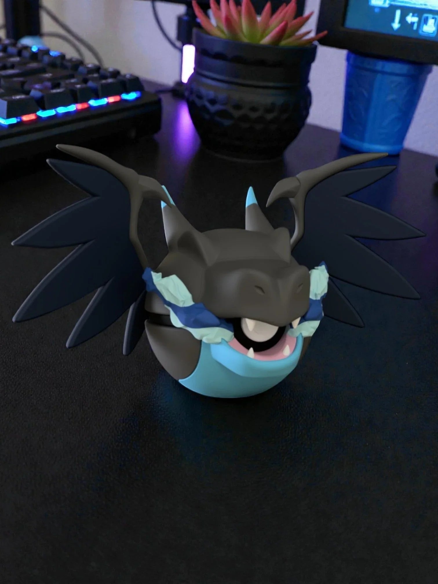 Pokeball edizione Mega Charizard X nera e blu, con ali e dettagli anime, su scrivania.
