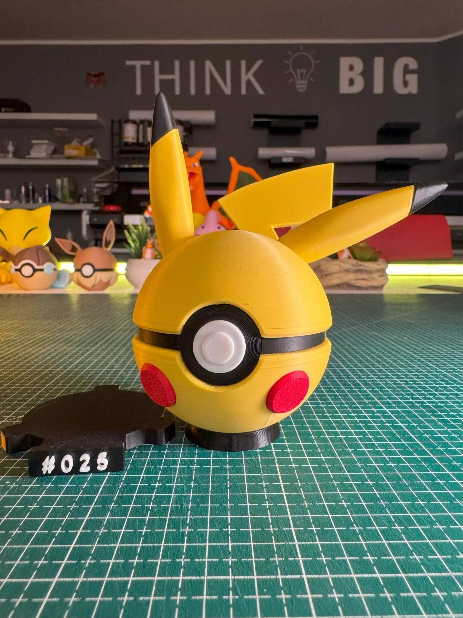 Pokeball gialla ispirata a Pikachu con orecchie e base numerata #025, su tavolo da lavoro.
