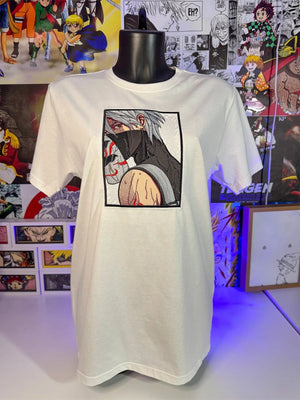 T-shirt bianca ricamata con personaggio anime mascherato, esposta davanti a sfondo di manga.