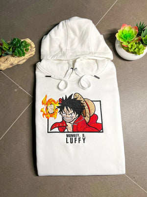 Felpa bianca ricamata con Monkey D. Luffy di One Piece, piegata su sfondo neutro, piante decorative ai lati.