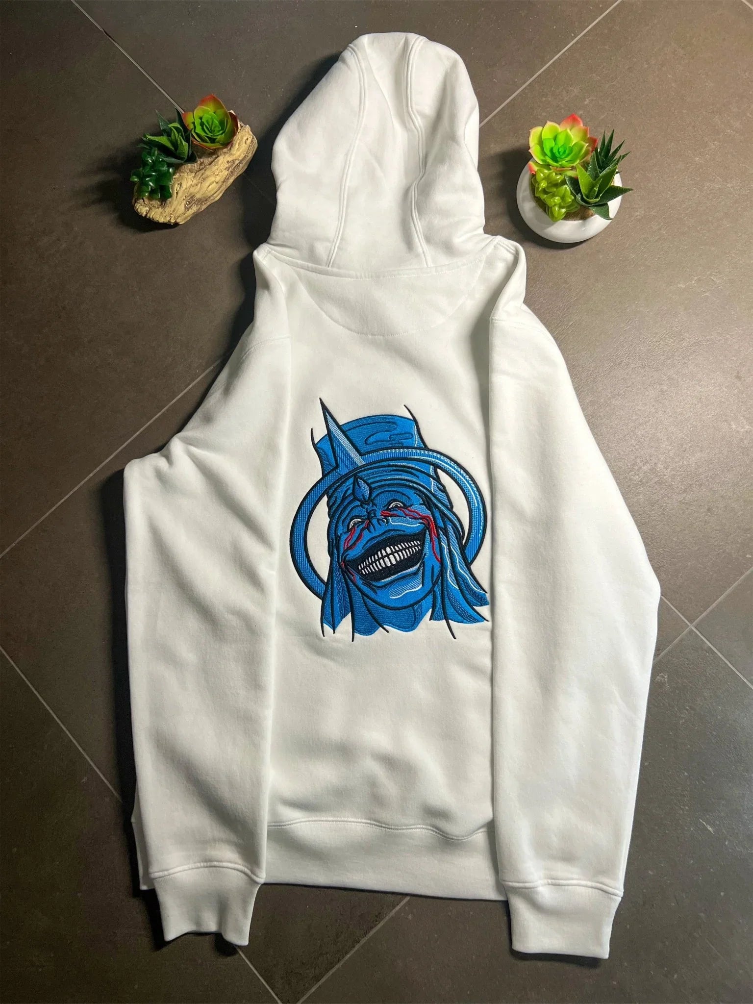 Felpa bianca con cappuccio, ricamo statua blu stile anime, fotografata su pavimento con piante grasse.