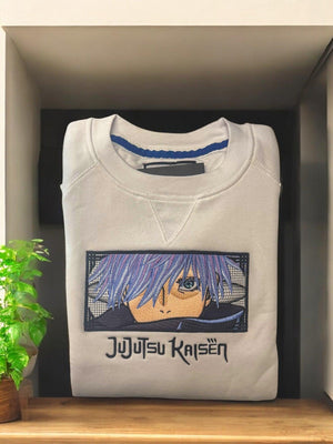 Felpa bianca ricamata con Gojo Satoru di Jujutsu Kaisen, piegata su scaffale.