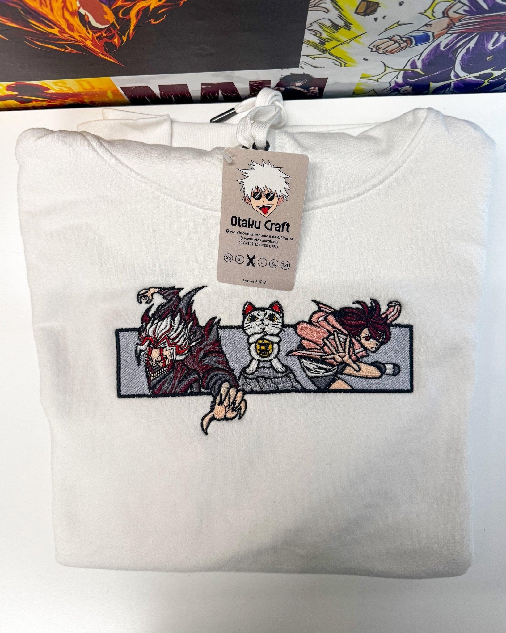 Felpa bianca ricamata con personaggi anime e gatto portafortuna, piegata su sfondo manga.