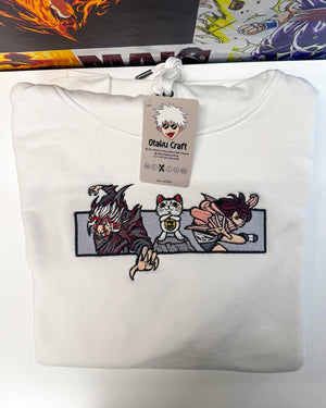 Felpa bianca ricamata con personaggi anime e gatto portafortuna, piegata su sfondo manga.