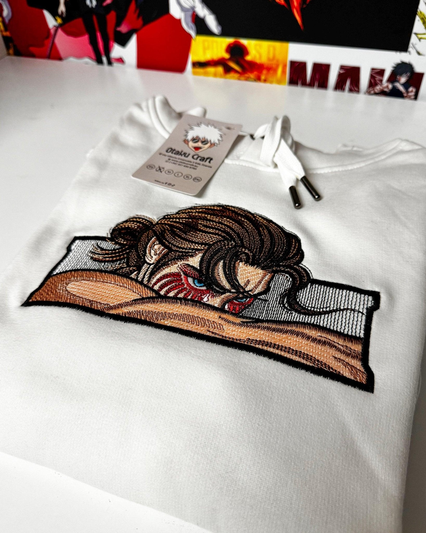 Felpa bianca ricamata Eren Yeager Attack on Titan, dettaglio anime e tag sullo sfondo.