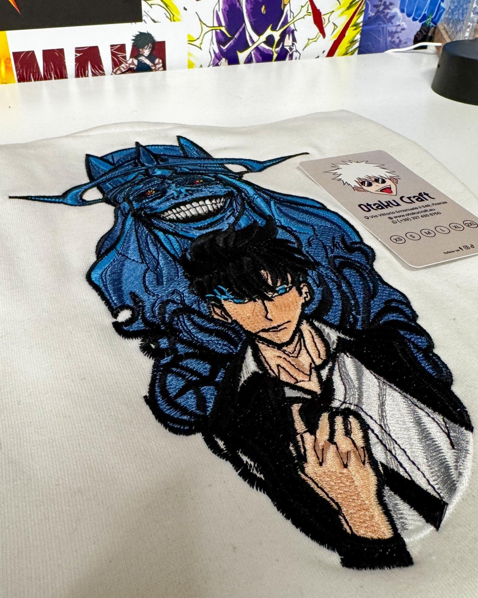Felpa bianca ricamata con personaggi anime Solo Leveling su sfondo di poster manga.