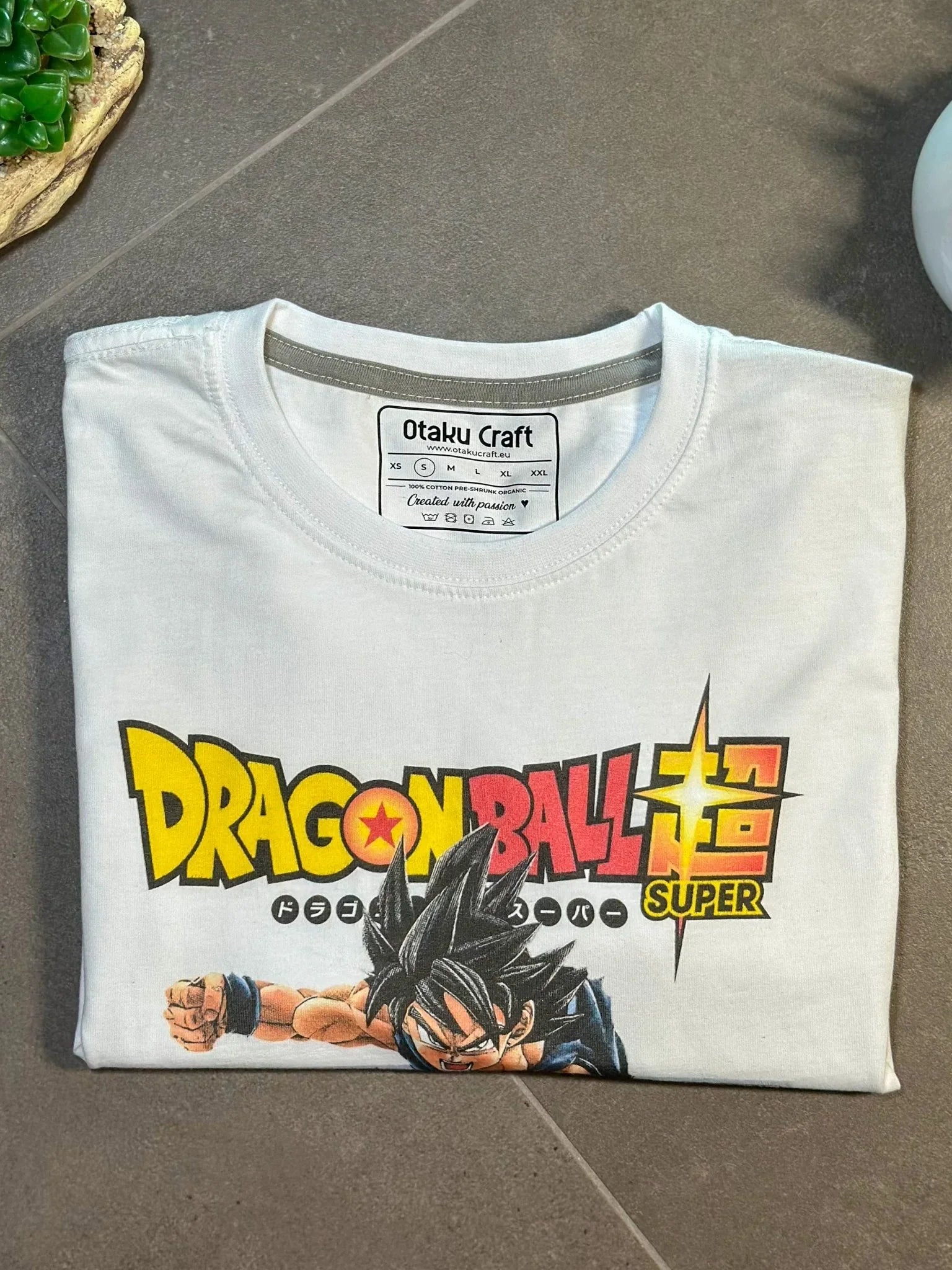 Maglietta bianca Dragon Ball Super con stampa Goku e logo colorato, formato piegato.