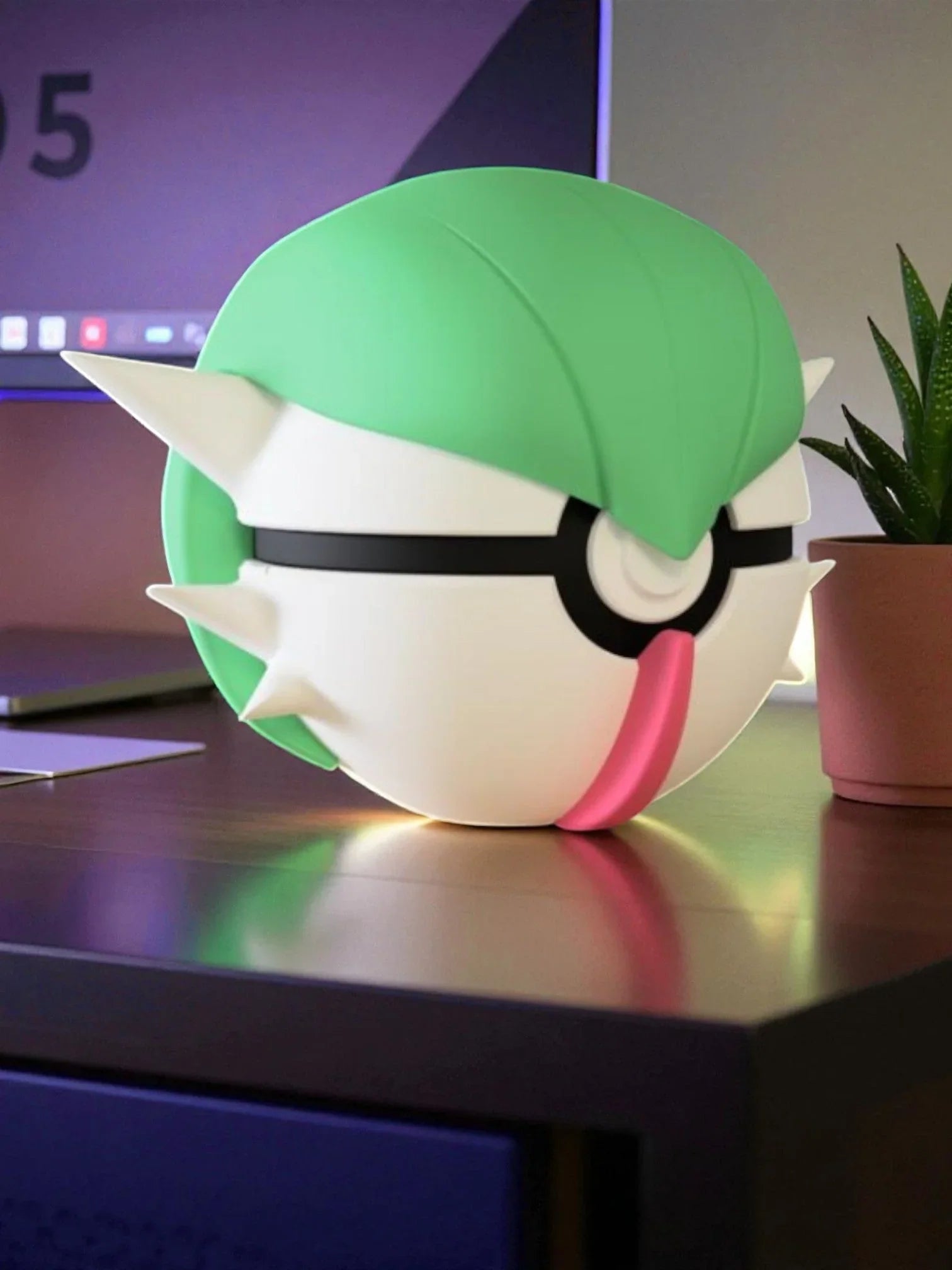 Pokeball ispirata a Gardevoir con dettagli verde e rosa su scrivania accanto a pianta