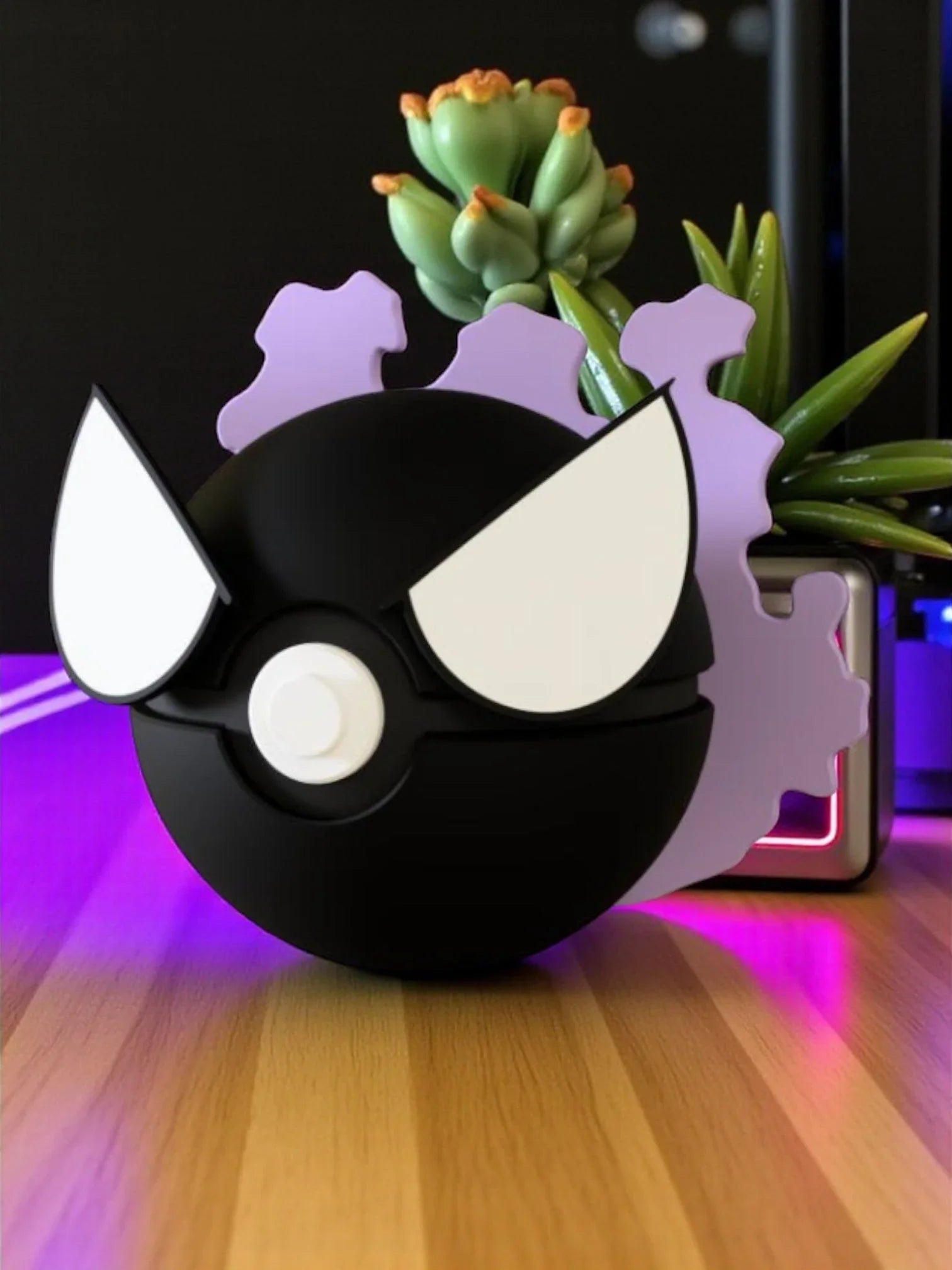 Pokeball a tema Gastly nero con occhi bianchi e dettagli viola su tavolo di legno, pianta sullo sfondo