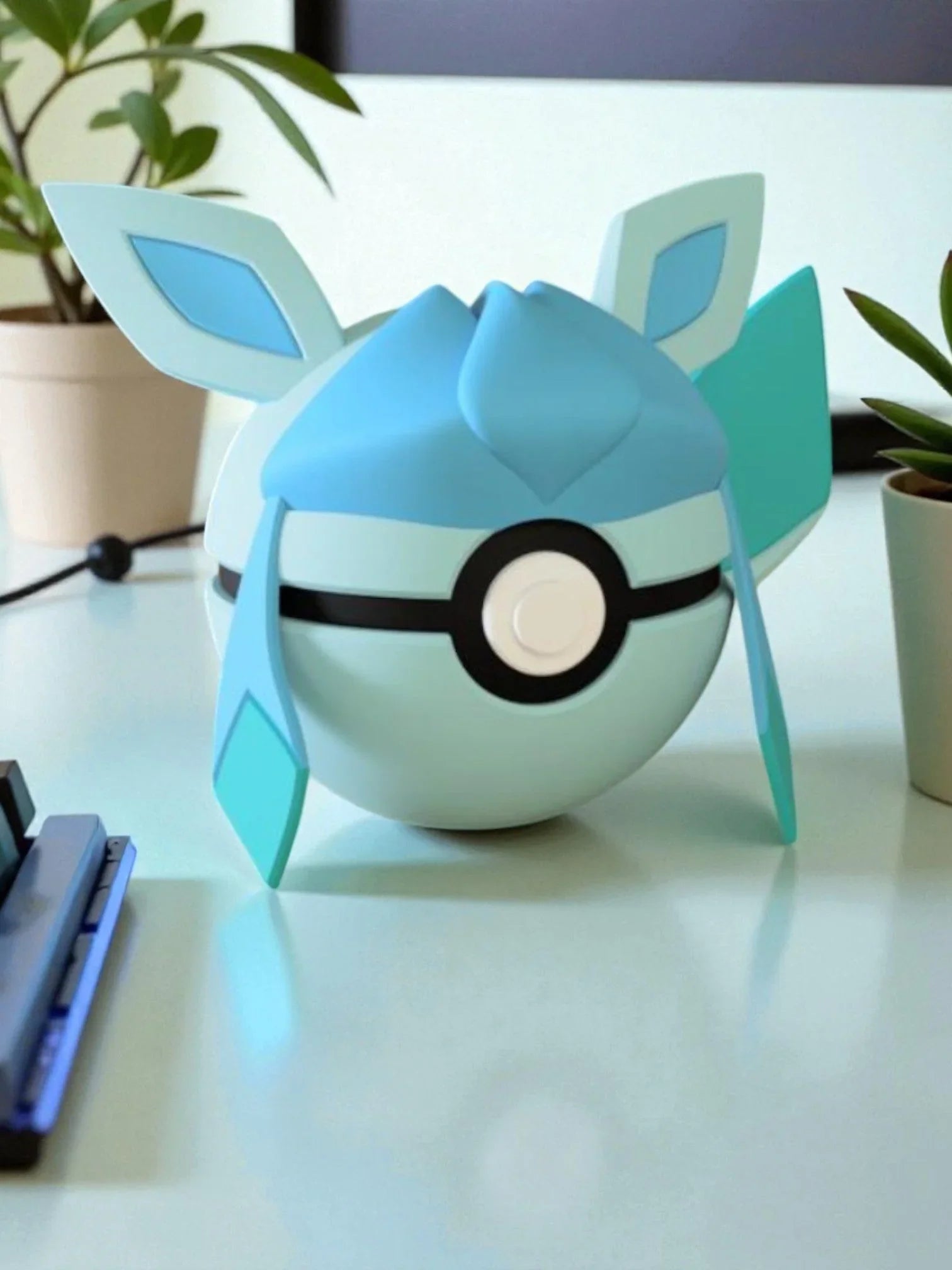Pokeball ispirata a Glaceon azzurra con orecchie stilizzate su scrivania, piante vicino